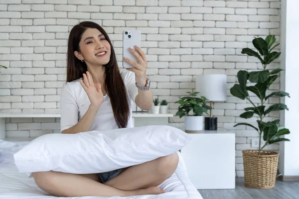 A casual day of a young woman on a bed in a bedroom. A young woman sitting with pillows on a bed and video call on smartphone