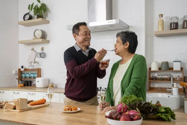 Happily Mature couple have spending time together in kitchen, husband Feeding tasting sausage to his wife, They smile and talk with happiness together