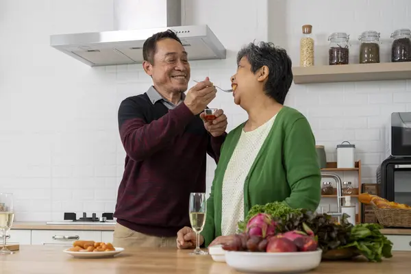 Happily Mature couple have spending time together in kitchen, husband Feeding tasting sausage to his wife, They smile and talk with happiness together