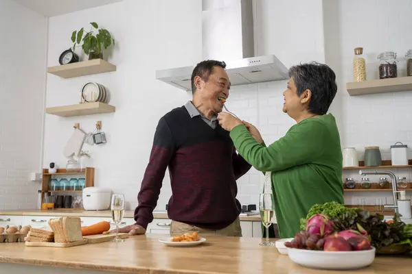 Happy healthy mature older family having breakfast at home. Wife feeding husband tasting sausage  in kitchen, embracing enjoying morning meal together, talk and laugh with fun