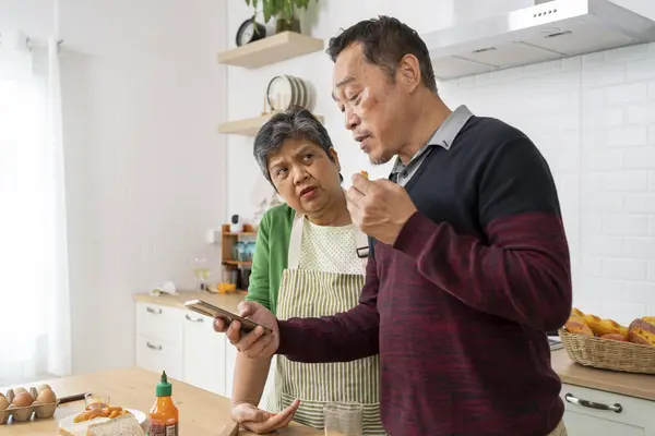 Happy healthy mature older family having breakfast at home. Wife and husband use smartphone during eating sandwich in kitchen together