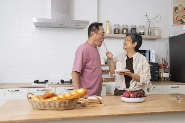 Asian mature couple eating dragon fruit in kitchen, Wife feeding tasting fresh fruit to husband, They are talking and smile with happiness together