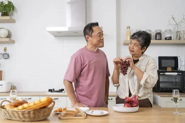 Asian mature couple playfully tease each other with grapes in kitchen, They talk and laugh with fun during eating fresh fruits with red grape, husband hugging wife with love