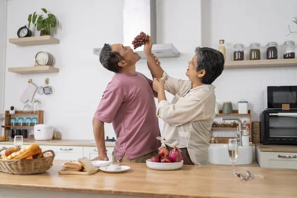 Asian mature couple playfully tease each other with grapes in kitchen, They talk and laugh with fun during eating fresh fruits with red grape, husband hugging wife with love