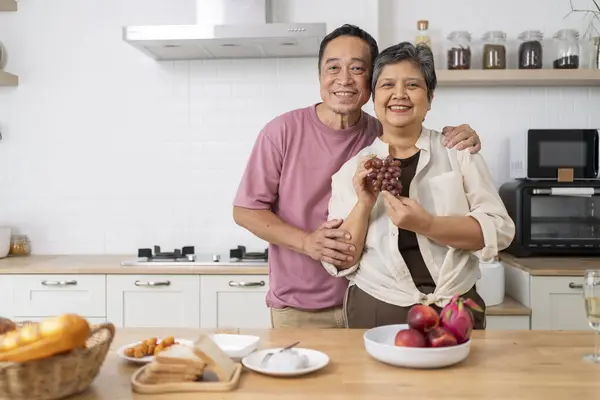 Portrait of Asian mature couple showing fresh fruits with red grape, husband hugging wife with love, They are looking at camera with smile