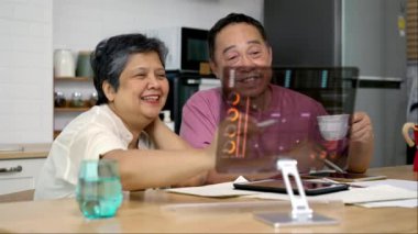 Orta büyüklükte el kamerası, Asyalı olgun çift bilgisayarla çalışmak için teknolojik aygıt kullanıyor, evde finans hakkında grafik tasarımı olan şeffaf monitör kullanıyorlar, tartışıyorlar ve başarılı olduklarından memnunlar.