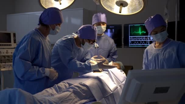 手持式枪法 专业的外科医生在手术室对床上麻醉的女病人进行手术切除 年轻的男性助手站在旁边 看护病人交出手术刀 — 图库视频影像