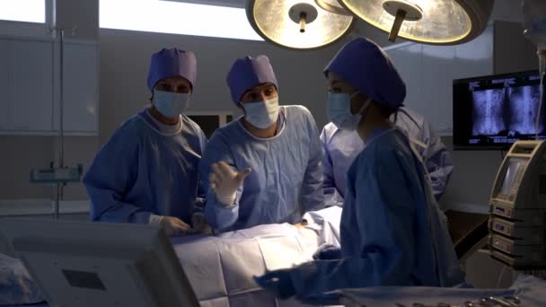 手持式拍摄 专业的外科医生小组在完成手术后进行讨论 他们看着监控器的控制 一起交谈 在手术室的床上麻醉的女病人 — 图库视频影像
