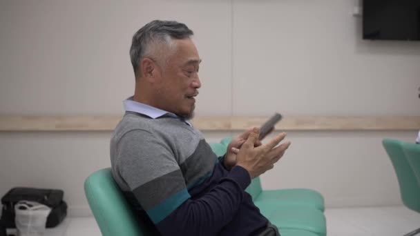 ハンドヘルドショット 選択的なフォーカス成熟した男性患者座って 病院のカウンターで支払いを待っている間に話すためにスマートフォンを使用 — ストック動画