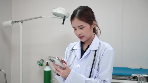 手持中枪 穿着实验室外套的年轻女医生站在考场 一边在智能手机屏幕上打滚一边休息 — 图库视频影像