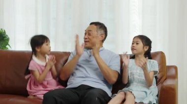 Orta boy, Asyalı emekli bir adam ve iki tatlı torunu oturma odasında kanepede oturuyor. El çırpmayı seviyorlar ve birlikte mutlu bir şekilde şarkı söylüyorlar. Aile sevgisinin sıcaklığı.