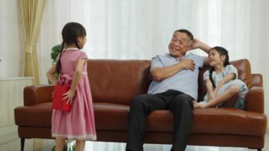 Orta boy fotoğraf, dedem ve tatlı kız kanepede oturup eğlenerek konuşuyorlar, küçük kız büyükbabasına sürpriz yaptığı için arkasına bir hediye kutusu saklıyor, onu sevgiyle kucaklıyor.