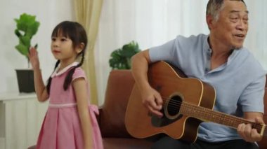 Orta boy, Asyalı emekli adam ve güzel torunu koltukta oturup gitar çalıyor ve şarkı söylüyor, küçük kız dans ediyor, müzikle mutlu bir şekilde eğleniyor, aile sevgisinin sıcaklığı.