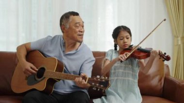 Orta boy fotoğraf, Asyalı büyükbaba gitar çalıyor ve şarkı söylüyor ve güzel torun kemanla oynuyor, müzikle eğleniyor, aile sevgisinin sıcaklığı...