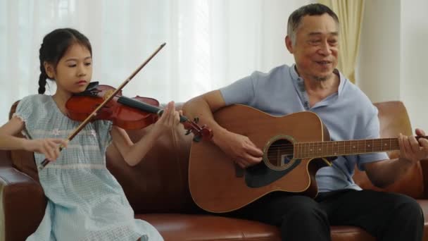 ハンドヘルドメディアショット アジアの引退した男 素敵な孫娘とソファに座って 祖父はバイオリンでギターを演奏し リトルガールは一緒に音楽を楽しむ 家族の愛の暖かさ — ストック動画