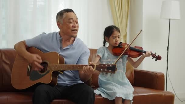 ハンドヘルドミディアムショット アジアの引退した男と素敵な孫娘ソファに座って 祖父はギターを演奏し 歌います リトルガールはバイオリンで遊びます 音楽と一緒に楽しむ 家族の愛の暖かさ — ストック動画