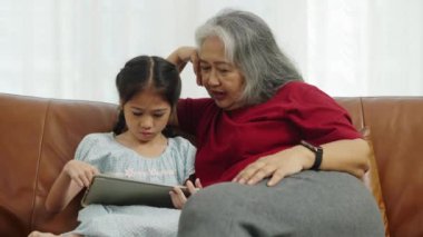 Görüntüler orta ölçekli, büyükanne ve iki torun kanepede oynuyorlar, tablet kullanıyorlar, oyun buluyorlar ve eğlenerek tartışıyorlar, oturma odasında mutlulukla gülüyorlar, aile sevgisinin sıcaklığıyla.