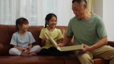 Görüntüler orta boy, iki Asyalı kardeş kanepede oturuyor, dedem elinde pizza kutusuyla içeri giriyor, evde birlikte lezzetli yiyecekler yedikleri için mutlu bir şekilde gülüyorlar.