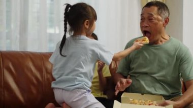 Görüntüler orta boy, iki Asyalı kardeş koltukta oturuyor, emekli adam kart kutusuyla geliyor, torun büyükbabaya pizza yediriyor, evde birlikte lezzetli yemekler yiyorlar.