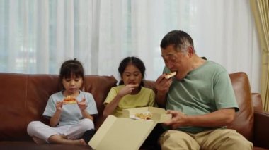 Görüntüler orta boy, iki Asyalı kardeş ve bir Emeklinin oturma odasında koltukta oturması, dedemle torunun evde lezzetli atıştırmalıklar yemeleri.