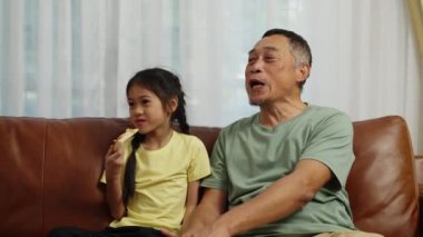 Görüntüler el kamerasıyla çekilmiş, iki Asyalı kardeş ve bir Emeklinin oturma odasında oturması, dedemle torunun evde lezzetli atıştırmalıklar yemeleri.