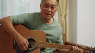 El kamerasıyla çekilmiş, emekli adam koltukta oturmuş akustik gitar çalıyor ve evde dinlenme odasında şarkı söylüyor.