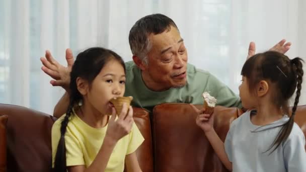 フッテージハンドヘルドミディアショット 2人のアジアの兄弟がソファーに座ってチョコレートアイスクリームコーンを食べ リビングルームで祖父と話すことを楽しむ — ストック動画