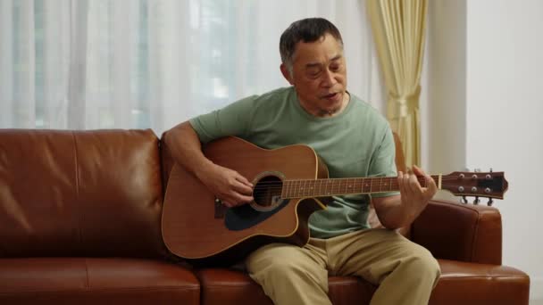 フッテージハンドヘルドミディアムショット 退職した男は ギターを演奏し 自宅のリビングルームでリラックスした時間で歌うソファに座って — ストック動画