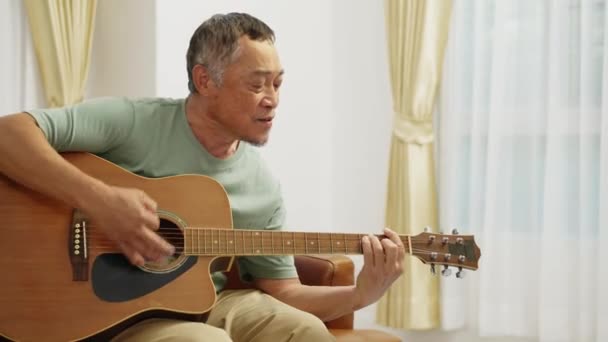 フッテージハンドヘルドミディアムショット アコースティックギターを演奏し 自宅のリビングルームでリラックスした時間を歌うソファに座っている退職した男 — ストック動画