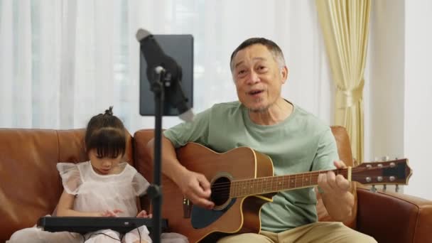フッテージハンドヘルドワイドショット アジアの引退した男はギターを弾き 2人の孫娘 小さな女の子は自宅のリビングルームのタブレットを通じてソーシャルネットワーク上で電気音楽キーボードショーを果たしています — ストック動画