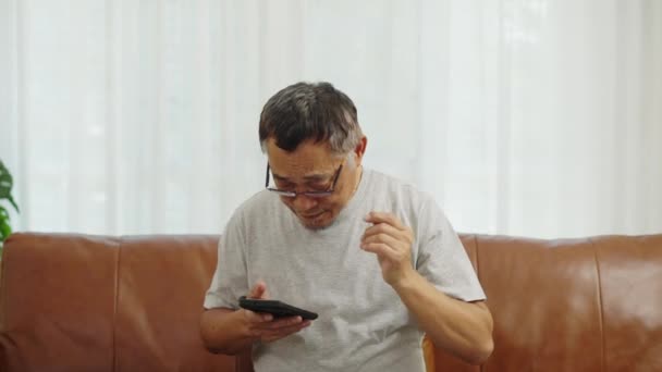 フッテージハンドヘルドミディアショット リラクゼーションのためにリビングルームにソファーに座って眼鏡をかぶっている幸せな成熟した男性 彼は手でスマートフォンを見て 眼鏡を離れて笑顔でカメラを見ています — ストック動画