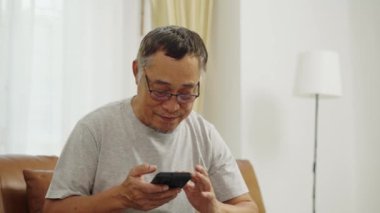 El kamerası görüntüsü orta boy, oturma odasında gözlüklü bir adam dinlenmek için koltukta oturuyor, elinde akıllı telefon ekranına kayıyor, sonra gözlüklerini çıkarıyor ve ileriye bakıyor.