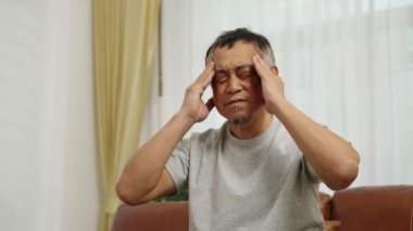 El kamerasıyla düşük açılı görüntü, oturma odasındaki koltukta oturan Asyalı emeklinin baş ağrısı yüzünden gözlerini kapatması baş ağrısını hafifletmek için her iki şakağına da masaj yapması.