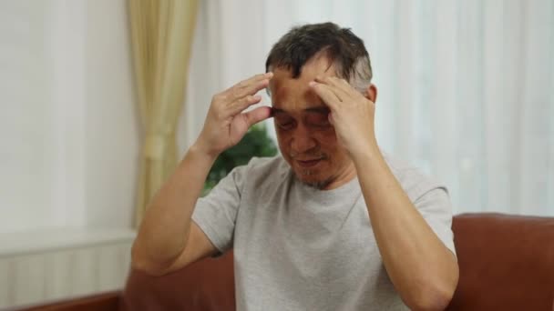 镜头紧紧抓住镜头 亚洲退休男子坐在客厅的沙发上 因头疼而闭上眼睛 他自己按摩头和额头以减轻头部疼痛 — 图库视频影像