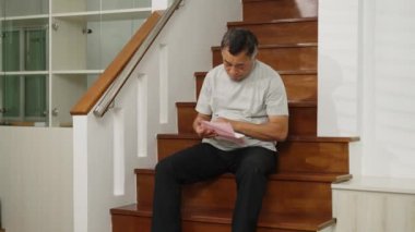 Görüntüler orta ölçekli, endişeli Asyalı emekli adam merdivenlerde oturuyor, ev giderleri için endişeleniyor, akıllı telefonu hesaplama ve kontrol için kullanıyor. Faturayı at, gerçek hayattaki finansal stres kavramı.