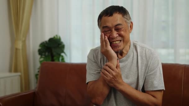 フッテージハンドヘルドミディアショット フロントビュー リビングルームにソファーに座っているアジアの退職男性 自分で歯痛を和らげるために頬をマッサージ — ストック動画