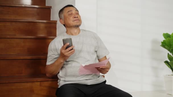 フッテージハンドヘルドミディアショット 懸念されたアジアの退職した男性は 家の費用を心配して階段に座ります 紙幣やスマートフォンを手に持つ現実世界の金融ストレスコンセプト — ストック動画