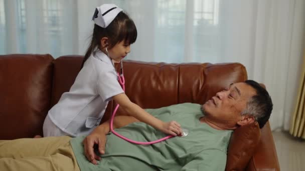 フッテージハンドヘルドミディアショット 看護師の制服を着て素敵な孫娘は 家でソファーに祖父の左胸に心臓の音を聞くスタチオスコープを使用して 彼らは一緒に楽しく話しています — ストック動画