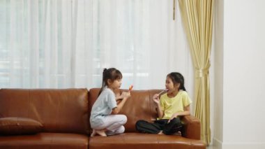 Görüntüler el kamerasıyla çekildi, iki tatlı kız kanepede oturuyor ve birlikte büyük lolipoplar yiyip eğleniyorlar, Asyalı kız kardeşler oturma odasında eğlenerek konuşuyorlar.