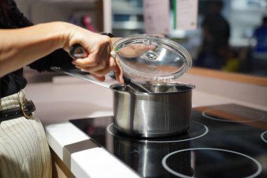 Seçici odak, mutfakta elektrikli ocakta lezzetli yemekler pişiren bir kadının elleri, elinde cam kap kapağı ve kepçeyle.