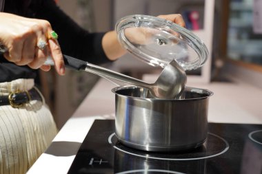 Seçici odak, mutfakta elektrikli ocakta lezzetli yemekler pişiren bir kadının elleri, elinde cam kap kapağı ve kepçeyle.