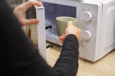 Kırpılmış görüntü, seçici odaklı kadın elleri mikrodalga fırını kullanarak mutfaktaki bardakta içecek ısıtıyor, boşluğu kopyalıyor.