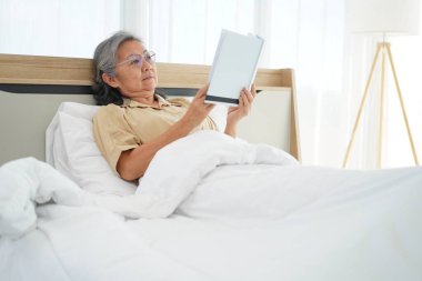 Gözlüklü, yatakta uzanıp kitap okuyan, yatak odasındaki yorganın altında dinlenen ve kitap okumaktan zevk alan mutlu olgun bir kadın. Boşluğu kopyala