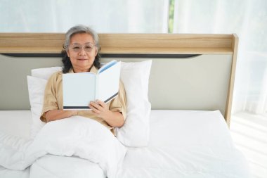 Gözlüklü, yatakta uzanıp kitap okuyan, yatak odasındaki yorganın altında dinlenen ve kitap okumaktan zevk alan mutlu olgun bir kadın. Boşluğu kopyala