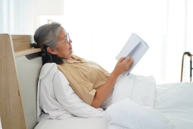 Gözlüklü olgun bir kadının yatakta yatıp kitap okuması yatak odasındaki yorganın altında dinlenmesi ve kitap okumanın keyfini çıkarması. Boşluğu kopyala
