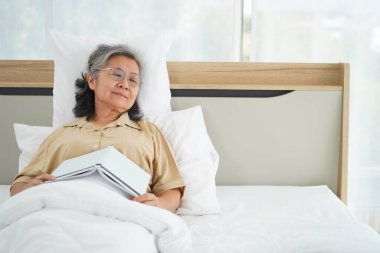 Gözlüklü Asyalı olgun bir kadın yatakta kitap okuduktan sonra uykuya dalıyor, yatak odasındaki yorganın altında dinleniyor, boşluğu kopyalıyor.