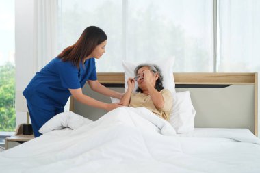Asyalı yetişkin gözlüğünü çıkar, genç hemşire asistanı yaşlı kadına yatakta yatmasına yardım et, bakıcı kadın battaniyeyle üstünü ört ve evinde rahat uyumasını sağla.