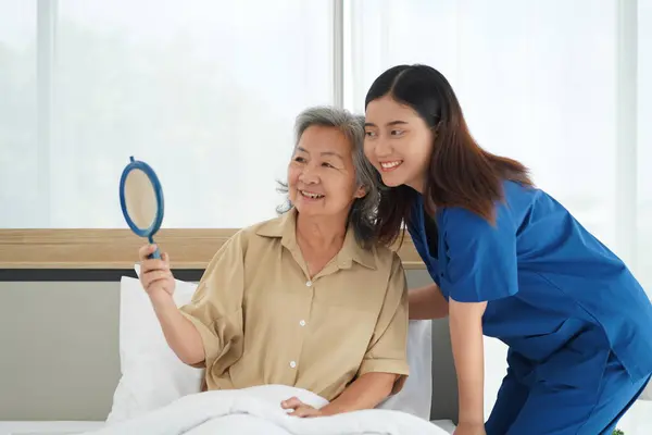 young nurse assistant female gently cares for an elderly woman, gently combing her hair on the bed, Senior woman looking a mirror with happy,  Caregiver providing comfort and reassurance at home