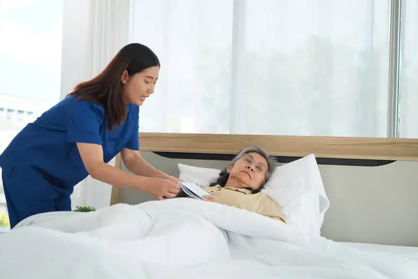 Young Nursing assistant remove a book during helping senior woman lying down in the bed, Caregiver female Help cover her with a blanket and make sure she sleeps comfortably at home