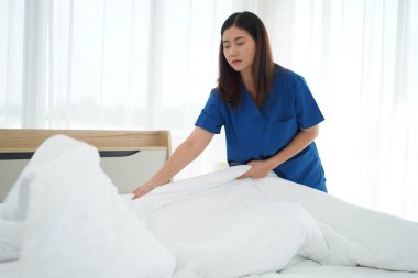Üniformalı genç bakıcı kadın yatak odasındaki yastıkları ve beyaz yorganı temizliyor ve örtüyordu. Yatma saatine hazırlanıyordu. Yaşlılara bakmak için günlük aktiviteler.